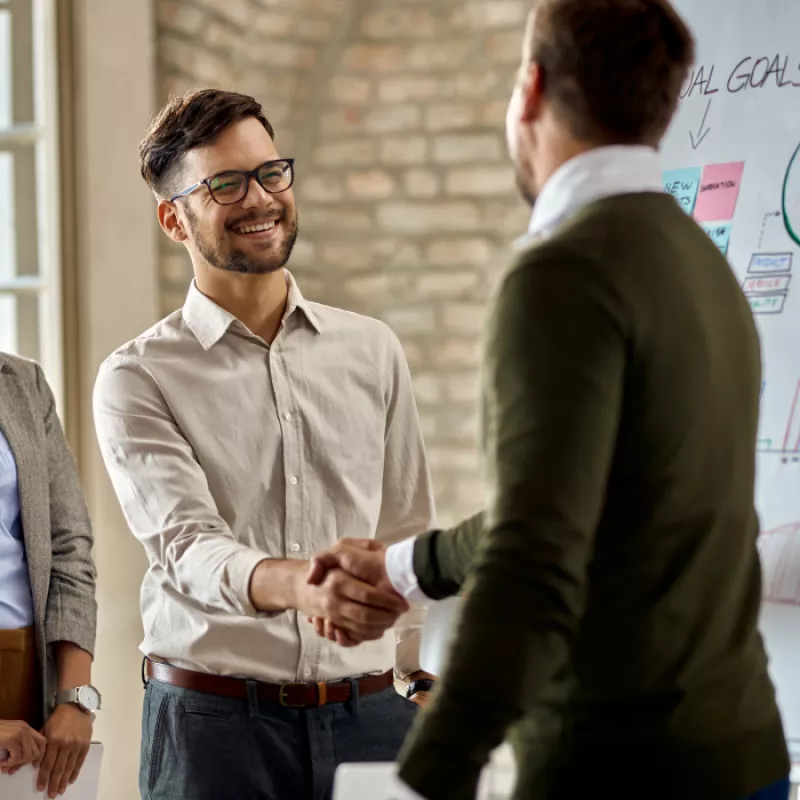 Two professionals engaged in a handshake, while a background board displays charts, conveying a positive and successful business interaction